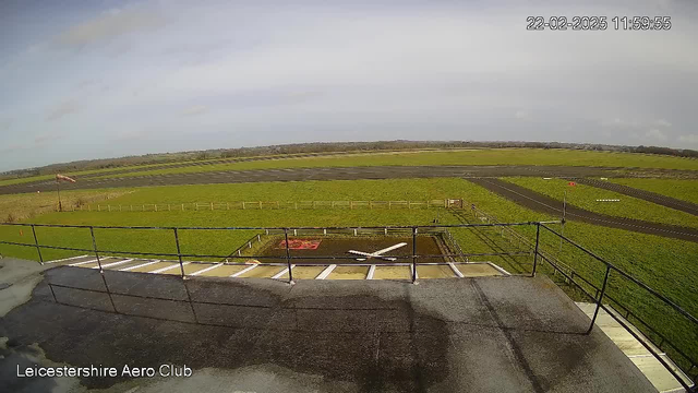 A view from a high vantage point at the Leicestershire Aero Club, showing a grassy landscape and a runway. The foreground features a black rooftop with white stripes, and a railing along the edge. Below, there is a red and white airplane symbol on the ground, surrounded by dirt. In the background, expansive green fields stretch out, with a dark runway running horizontally across the scene, and a light sky with scattered clouds overhead.