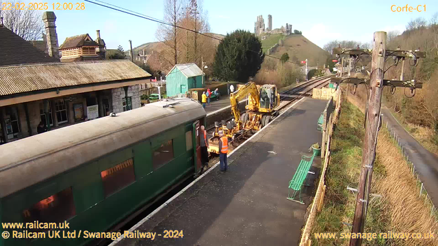 A green train is parked at a railway station platform. Two workers in orange safety vests are near the train, one holding equipment while a yellow construction vehicle is positioned close to the train tracks. In the background, there are several trees and a hill with a castle ruins at the top. The sky is clear and sunny, and there are a few people walking around the station area. Green benches are visible along the platform, and a telephone pole with wires stands on the right side of the image.