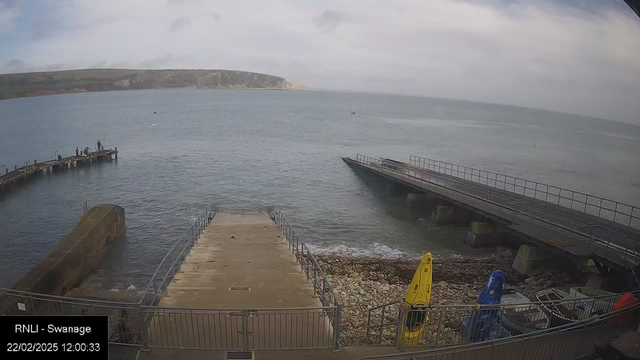 A view of a calm sea under a cloudy sky, with a rocky shore and a visible pier on the left. At the end of the pier, there are two people fishing. There are two boats moored nearby, one yellow and one blue, positioned on the shore next to the pier. The area is surrounded by cliffs in the background, suggesting a coastal landscape. The image appears to be taken during the day.