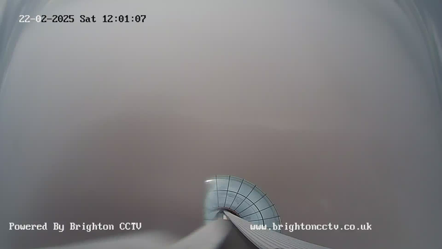 A foggy scene captured by a webcam, with the view primarily obscured by grey mist. In the foreground, a curved glass structure with a grid pattern is partially visible. The time stamp in the upper left corner reads "22-02-2025 Sat 12:01:07." At the bottom left, the text "Powered By Brighton CCTV" is displayed, along with a website link on the bottom right.