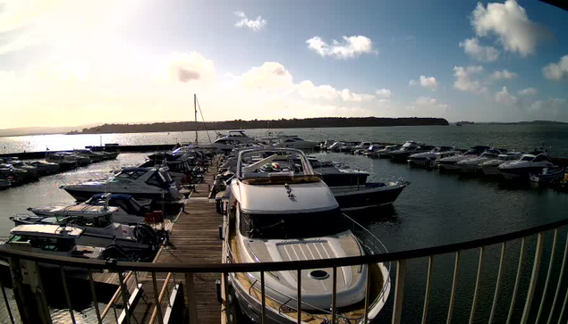 A scenic view of a marina on a clear day. Numerous boats are docked on the water, with a mix of small and larger vessels visible. The sky is partly cloudy, and the sunlight reflects off the water's surface, creating a shimmering effect. In the background, there are lush green hills. A wooden dock extends into the marina, and a railing is visible in the foreground, adding depth to the scene.