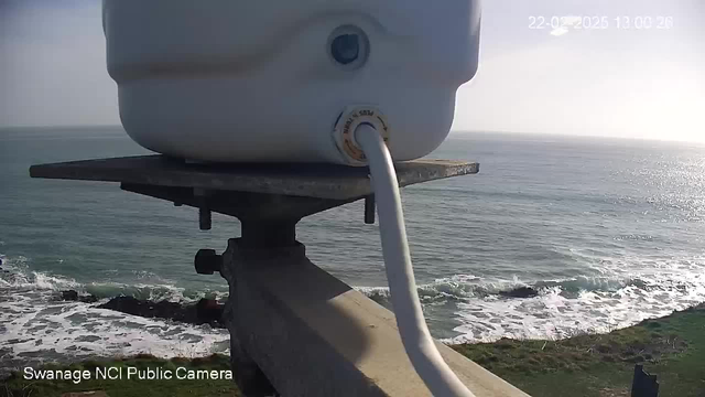 A white container is mounted on a metal platform, with a cable extending from it. In the background, the ocean is visible with gentle waves and a clear sky. The shoreline is visible in the foreground, with rocky formations and some greenery. The image is taken during the daytime, suggesting bright lighting conditions. A timestamp indicating the date and time appears in the upper right corner.