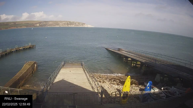 A seaside view featuring calm blue waters under a partly cloudy sky. In the foreground, there are two wooden piers extending into the water, with one pier wider and located on the right. To the left, a rocky shore is visible with some stones and a small section of a wall. Two yellow and blue kayaks are placed on the shore near the wooden structures. In the background, there are green hills and cliffs along the coastline, with a small white sailboat spotted on the water.