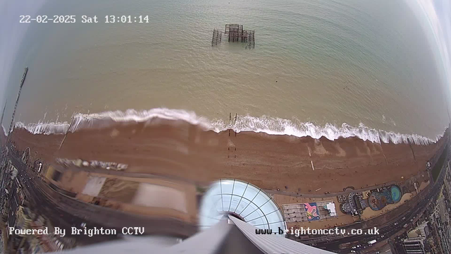 Aerial view of a beach with brown sand and gentle waves lapping at the shore. In the water, there is a wooden pier structure partially submerged. To the right, there are scattered buildings and a swimming pool, with a brightly colored pattern visible on the rooftop of one of the buildings. The sky is overcast, and the scene is captured by a webcam. A timestamp indicates the date as February 22, 2025, at 13:01:14.