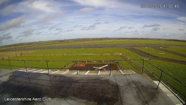 A view from a webcam at the Leicestershire Aero Club, showing an expansive green field and an airstrip. In the foreground, there's a railing and a wet surface. The runway is visible, marked with white lines. The sky is partly cloudy with patches of blue and some scattered clouds. In the distance, there are more fields and trees, with a small red flag appearing at the edge of the field.