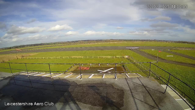 A wide view of an airfield under a cloudy sky. In the foreground, there is a railing indicating a viewing platform. Below the platform, there is a helipad marked with a white cross and adjacent to it, a grassy area. Beyond the helipad, stretches a large field covered in green grass, bordered by a wooden fence. In the distance, there are landing and takeoff runways made of asphalt. A small airplane is visible on the runway towards the right side of the image.