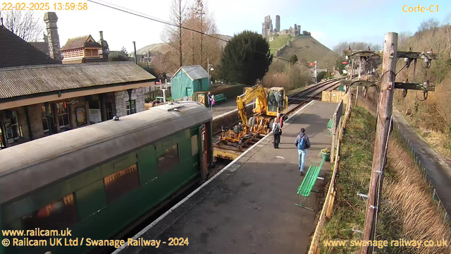 A train station scene with a green vintage train on the left. A construction vehicle is positioned next to the tracks, and two people, one with a backpack and the other with a small bag, are walking on the platform. In the background, there are trees and a small green shed, with the remnants of a castle visible on a hill in the distance. The sky is clear and bright, indicating daylight.