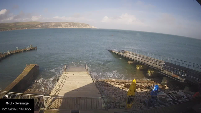 A coastal scene showing wooden walkways leading down to the water's edge at Swanage. The view features a gentle wave action on the surface of the blue sea, with rocky shoreline visible on the left. Two brightly colored kayaks, one yellow and one blue, are positioned near the walkway, alongside scattered pebbles and rocks. In the background, a green hill rises with a cliff face further along the horizon, under a partly cloudy sky.