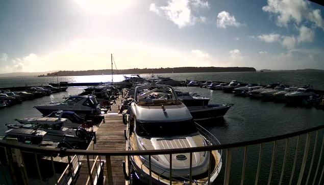 A view of a marina filled with multiple boats docked by a wooden pier. In the foreground, a large, white motorboat is prominently positioned. The scene is illuminated by bright sunlight, casting reflections on the water, which gently ripples around the boats. In the background, green hills can be seen on the horizon, under a partly cloudy sky.