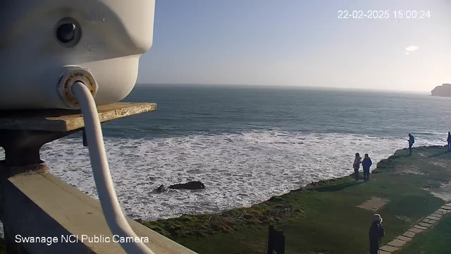 A coastal scene captured from a webcam, showing a calm sea under a clear sky. In the foreground, there is a white structure with a cable extending from it, possibly part of the camera setup. The background features gentle waves lapping against the shore and a patch of grass. Several people are standing along the coastline, observing the view. The date and time displayed in the corner indicate it is February 22, 2025, at 15:00:24.