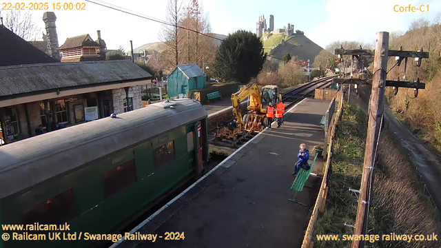 A train, partially visible, sits at a railway station platform. Two construction workers in bright orange vests operate machinery on the tracks. A young child sits on a green bench nearby, looking at a handheld device. In the background, there is a quaint building with a slanted roof, and the ruins of a castle are visible on a hill. The scene is set on a clear day with trees lining the edges of the platform.