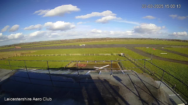 A view from a webcam at Leicestershire Aero Club, capturing a large green grassy field under a bright blue sky with some white clouds scattered. In the foreground, a solid structure with a cross marking on the surface suggests a helipad or landing area. A small aircraft is on the runway, moving towards the left. Near the runway, there is a wooden fence, and in the distance, there are patches of land with trees lining the horizon. A wind sock is visible, indicating wind direction.