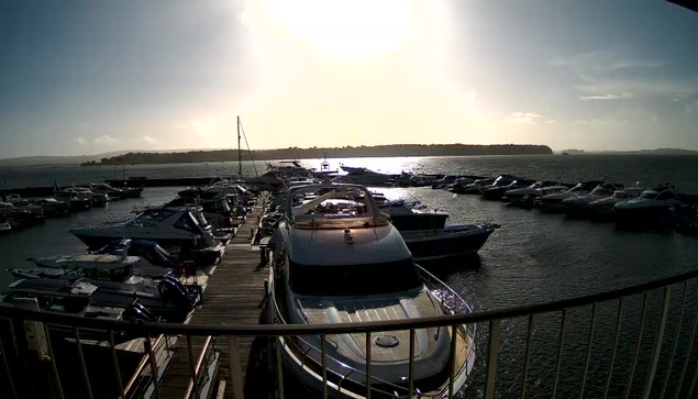 A marina with numerous boats docked along a wooden pier. The scene is illuminated by bright sunlight reflecting off the water, creating a shimmering effect. In the background, a stretch of land is visible across the water, and the sky is mostly clear with a few clouds. The boats vary in size and shape, some are larger yachts while others are smaller vessels. The atmosphere appears calm and peaceful.