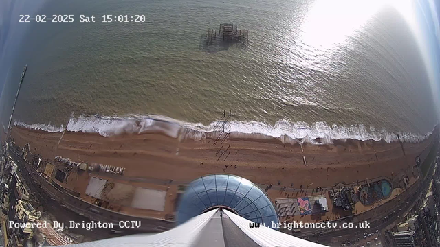 Aerial view of a sandy beach with people walking on it, waves gently lapping at the shore. In the water, the remains of a pier are visible. The coastline features a promenade lined with buildings, including an amusement park with colorful rides. The image is captured from a high vantage point, showing the blue ocean under a bright sky.