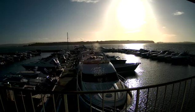 A marina scene is depicted with several boats docked along a wooden pier. The sun shines brightly in the sky, reflecting off the water's surface, creating a shimmering effect. In the background, a hilly shoreline is visible, with a few distant boats on the water. The overall atmosphere is peaceful and sunny.