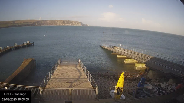 A view of a coastal scene featuring calm turquoise waters under a clear blue sky. In the foreground, there is a wooden pier extending into the water, with a stairway leading down to the shore. To the right, several small boats are moored, including a bright yellow craft and a blue one. The rocky shoreline is visible, and in the background, there are cliffs bordered by green hills. The time displayed is 16:00 on February 22, 2025.