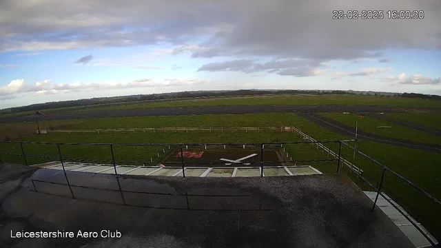 A view from a webcam positioned at the Leicestershire Aero Club shows a grassy airfield under a partly cloudy sky. In the foreground, there is a large X marking the center of a landing area. Surrounding the landing area, a fence defines the perimeter, and in the distance, a runway runs horizontally across the image. The ground is mostly green, with patches of earth and asphalt visible. The time and date displayed on the image show 16:00:30 on February 22, 2025.