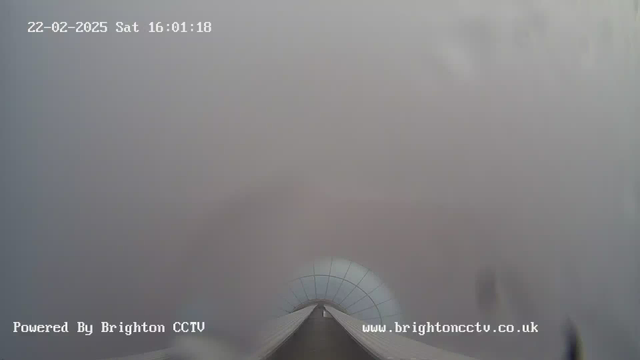 A view looking down a long, narrow pathway that appears to lead towards a structure with a dome shape at the end. The image is mostly obscured by fog or mist, making it difficult to see details beyond a few meters. The scene suggests a cloudy or overcast day. At the bottom, there are details indicating it is powered by Brighton CCTV, along with a timestamp displaying the date and time.