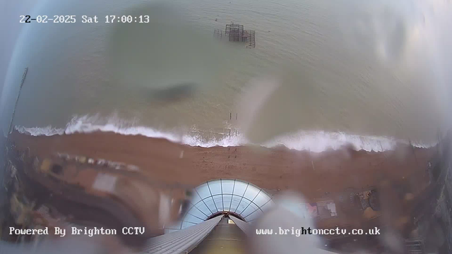 A view from a webcam positioned above a beach, showing the shoreline with waves gently lapping at the sand. The scene captures a clear sky and water, along with a partially visible structure, possibly a pier, in the water. At the bottom of the image, part of a building structure with curved glass can be seen, suggesting the viewpoint is from a height. The timestamp and logo are visible in the corners of the image.