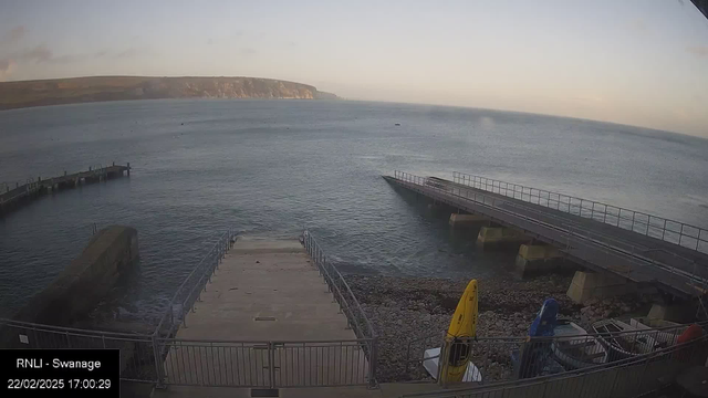 A view of a shoreline with rocky beach and water. In the foreground, there is a concrete ramp leading down to the water, bordered by a metal railing. To the right, two piers extend into the sea, one being shorter than the other. Several colorful kayaks are positioned on the shore near the ramp. The background features a coastline with hills and cliffs under a light sky, indicating either sunrise or sunset. The overall scene suggests a calm marine environment.