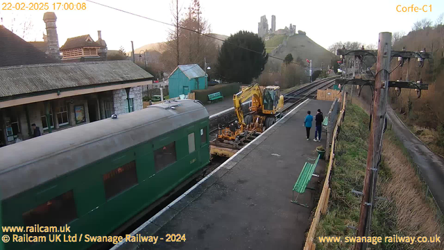 A green train carriage is positioned on the left side of the image, partially obstructing the view of the railway station platform. To the right, there is a yellow construction vehicle working on the tracks. Two people, one wearing a blue jacket and the other in black, walk along the platform. In the background, there are trees and a hill with ruins atop it. The date and time are displayed in the top left corner, and there are benches and a small building in the station area.