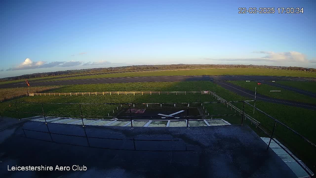 A clear sky with a few scattered clouds is visible above an open green field. In the foreground, there is a marked runway with a light-colored strip, and a white static airplane silhouette is positioned on the ground. The field is bordered by a wooden fence. The scene is peaceful, showing no visible activity or movement.