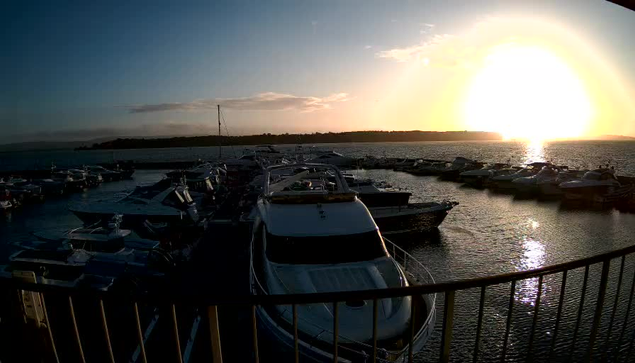 A scenic view of a marina at sunset, featuring numerous boats moored in calm water. The sun is setting on the horizon, casting a warm glow across the scene, with reflections shimmering on the water's surface. The background has silhouette outlines of land and a sky with soft clouds. A railing is visible in the foreground, framing the view.