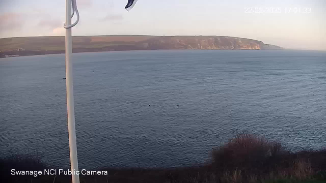 A view of the ocean with calm waters and small boats scattered across the surface. In the background, high cliffs rise against a clear sky. A flagpole is positioned on the left side of the image, with the flag gently waving. The horizon is visible where the sea meets the sky, and the overall scene conveys a tranquil coastal landscape.