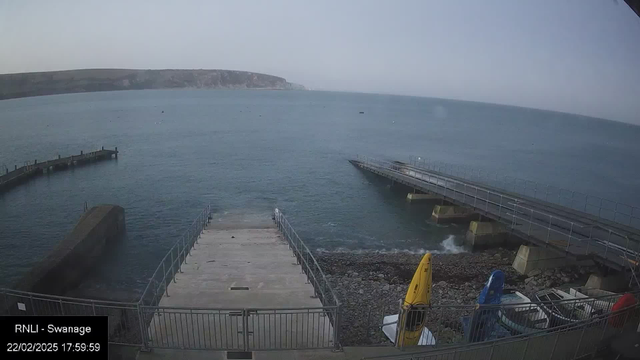 A view of a quiet coastal area with a stone jetty extending into the water on the left side. The foreground shows a wide ramp leading down to the water, with a railing on either side. To the right, there are several boats, including a yellow and a blue kayak. The water is calm and has a light ripple, with a distant shoreline visible in the background. The sky is overcast and gray, suggesting a cloudy day. 

Date and time are displayed at the bottom left corner: "22/02/2025 17:59:59 RNLI - Swanage."