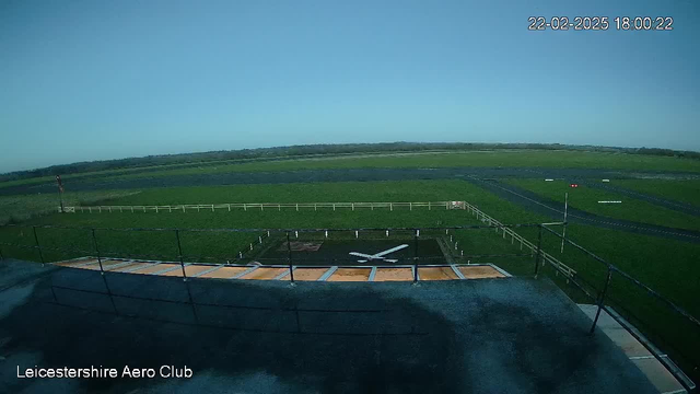 A wide view of an aerodrome under a clear blue sky. The foreground shows a flat roof with multiple rows of grayish panels and a large white 'X' marking on the ground. In the background, an expansive green field stretches across the image, bordered by a wooden fence. The horizon is distant with patches of trees and possibly more fields. A red flag and other markings are visible along the runway area. The overall scene conveys a calm, open outdoor space often associated with aviation activities.