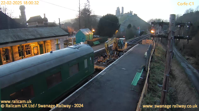 An image of a railway station during dusk. To the left, there is a green vintage-style train parked on the platform. A yellow construction vehicle is seen on the tracks next to the train, with tools and machinery for maintenance. The station building has a slate roof and features large windows, with a sign indicating "Corfe Castle." In the background, the silhouette of Corfe Castle is visible atop a hill, with trees and a few lights around the area. The scene is quiet, suggesting a peaceful atmosphere as day transitions into night.