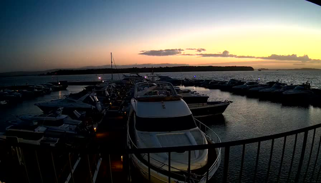 A waterfront scene at dusk featuring numerous boats docked in a marina. The boats are illuminated by soft lights, reflecting on the calm water. The sky transitions from light to dark, with hints of orange and purple hues as the sun sets. In the background, silhouettes of distant land and gentle clouds are visible. A railing frames the lower part of the image.