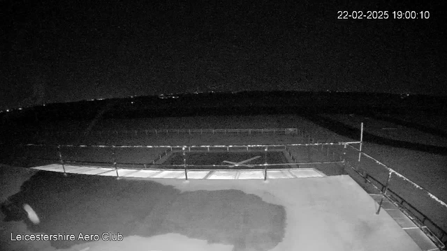 A dark webcam image showing a flat surface with a faint shadow on the left side. There is a low railing along the bottom, with faint horizontal lines visible. The background is mostly dark with a few distant lights suggesting a night scene, likely outdoors. The image is labeled with the date and time in the top right corner, indicating it was taken on February 22, 2025, at 19:00:10.