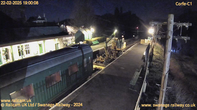 A nighttime image of a railway station. The scene is illuminated by several lights. In the foreground, there is a green train carriage on the left and a yellow construction vehicle positioned on the right. The platform is empty, with benches visible along the left side. The station building, with large windows and green accents, is located in the background. There are also trees and bushes in the surrounding area, adding to the nighttime atmosphere.