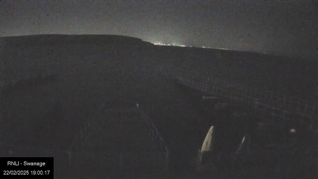 A dark, nighttime scene showing a coastline. The silhouette of a hill is visible in the background, while faint lights from a distant town can be seen on the horizon. In the foreground, there are outlines of structures, possibly boats or equipment, with a wooden boardwalk or pier extending across the bottom of the image. The atmosphere is quiet and dimly lit.