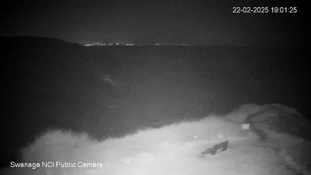 A nighttime view of a beach with low visibility. The dark water is gently lapping against the shore, which is partially obscured by foam. In the foreground, there is a wooden bench facing the water. A small sign or object is visible near the bench. In the background, faint lights from a distant coastal area can be seen against the horizon. The overall scene is dimly lit, with a sense of calmness in the atmosphere.