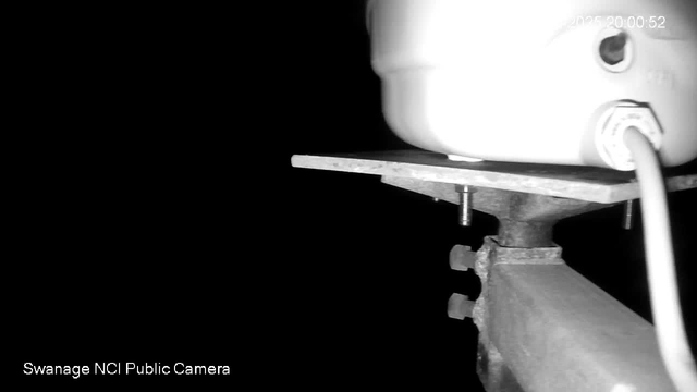 A dark image showing the lower portion of a white cylindrical object mounted on a flat surface. The object has a wire connected to it, and there are screws visible holding the surface in place. The background is mostly black with minimal light. The date and time are displayed in the top right corner. Text at the bottom left indicates "Swanage NCI Public Camera."