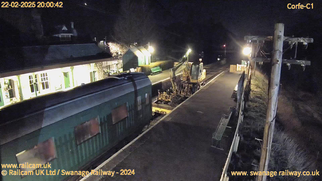 A dimly lit train station scene at night. In the foreground, a green train car is parked on the tracks. To the right, there is heavy machinery, possibly for maintenance, positioned on the platform. The platform features a few benches and some fencing. There are lit windows in the station building, revealing a warm yellow glow, while a few trees are silhouetted against the night sky. A streetlight illuminates part of the scene, casting shadows on the ground. The overall atmosphere is quiet and still, typical of a nighttime setting.