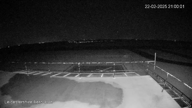 A dark night scene viewed from a webcam at Leicestershire Aero Club. The foreground features a flat surface with a faint wet area. A railing is visible in the lower part of the image, partially obstructing a view of the horizon where distant lights can be seen. The sky is mostly dark with a hint of light from the horizon, indicating a sense of vastness and tranquility. The timestamp shows the date and time as February 22, 2025, at 21:00:01.