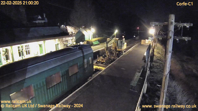 A nighttime view of a railway station platform. To the left, there's a green train parked, slightly obscured. In the center, a yellow construction vehicle is actively working. The platform is illuminated by yellow lights, with a couple of benches visible. In the background, there is a small building with lit windows, and a hint of greenery can be seen. The scene conveys a quiet, work-in-progress atmosphere typical of a railway setting at night.