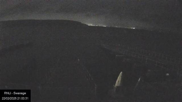 A dark image captured by a webcam, likely at night. The foreground features indistinct shapes that may represent boats or equipment, with a slight silhouette of a shoreline in the background. Faint lights are visible in the distance, suggesting a town or city across the water. The overall atmosphere is moody and shadowy, with limited visibility.