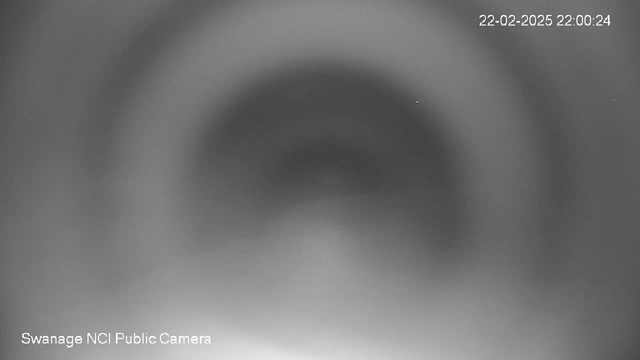 A mostly blurred black and white image featuring concentric circular patterns that fade towards the center. The background appears gray with a gradient effect. In the top right corner, there is a timestamp showing "22-02-2025 22:00:24," and in the bottom left corner, text reads "Swanage NCI Public Camera." The overall image lacks distinct objects or clear details.