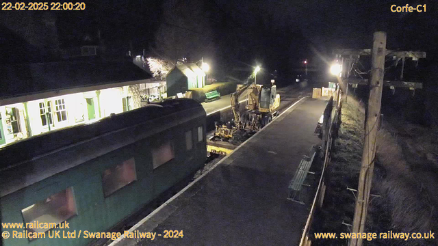 A dimly lit scene at a railway station at night. A green train carriage is positioned in the foreground. To the right, there is a yellow construction vehicle parked on the platform. The platform is empty, with a few benches and a small signal pole visible. In the background, a building with lit windows can be seen, along with a green bench facing the tracks. The surrounding area is mostly dark, contributing to the nighttime atmosphere.