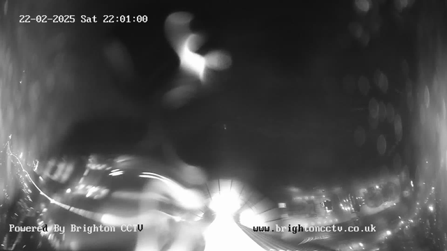 A black and white webcam image showing a blurred view of a roadway, with bright lights appearing at the center, indicating potential vehicle headlights. The date and time display at the top shows "22-02-2025 Sat 22:01:00." The lower section contains text "Powered By Brighton CCTV" and a website URL partially visible. The overall image has a hazy and unclear quality, suggesting low visibility conditions.