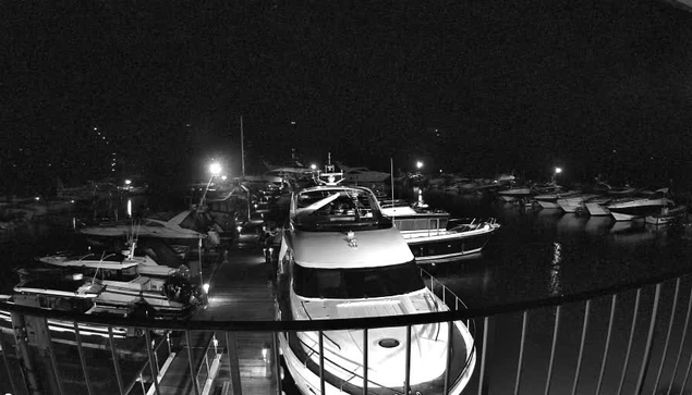 A dark waterfront scene featuring several boats docked in a marina at night. The foreground shows a large white yacht with its upper deck visible. Various smaller boats are moored beside it. Dim lights from the boats and the dock reflect on the water, creating soft glows in the dark setting. A railing is visible in the foreground, framing the view of the marina.