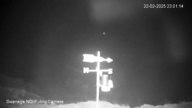 A black and white image shows a weather vane atop a pole, indicating north and south directions with visible arrows. The background is dark with unclear elements, suggesting night time conditions. A small light is visible in the top right corner of the image, possibly a star or other distant light source. The date and time are displayed in the top right corner: 22-02-2025 at 23:01:14. In the lower left corner, it says "Swanage NCI Public Camera."