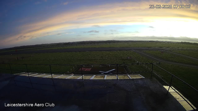 A view from a webcam at Leicestershire Aero Club, showing a grassy airfield under a pastel-colored sky at sunrise. In the foreground, there is a small model airplane on a patch of grass, surrounded by a fence. The runway is visible in the distance, along with a few scattered clouds in the sky. The date and time are displayed in the top right corner of the image.