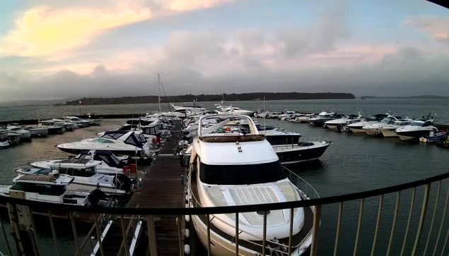 A marina scene with numerous boats docked in calm waters. In the foreground, several white and blue boats are visible, with one large white yacht prominently occupying the center. The background features more boats of various sizes, with a cloudy sky overhead transitioning from gray to a soft pink hue at the horizon. The marina is bordered by wooden docks and a railing, adding depth to the view.