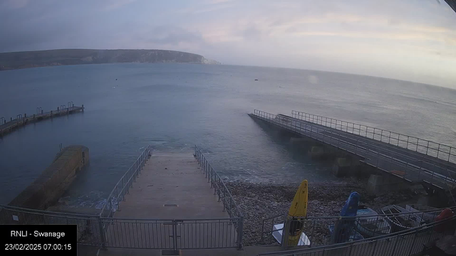 A view of a calm sea at dawn, with gentle waves lapping against a stone pier. The pier is lined by railings and extends into the water, while a smaller wooden dock is to the left. On the right side, there are two yellow kayaks and other boats secured on the shore, surrounded by pebbles. In the background, the coastline rises into a green, hilly area under a partly cloudy sky. The time and date displayed indicate it is early morning on February 23, 2025.