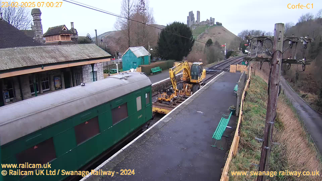 A green train carriage is parked on a railway platform, with a yellow construction vehicle next to it. In the background, there is a hill with ruins of a castle. The platform has benches and a few trees, with a building featuring a slate roof visible to the left. Overcast sky with a subtle light. Electric poles and wires are also present in the scene.